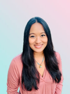 Ginny Dolores wears a pink shirt and a blue necklace against a pink and blue background.