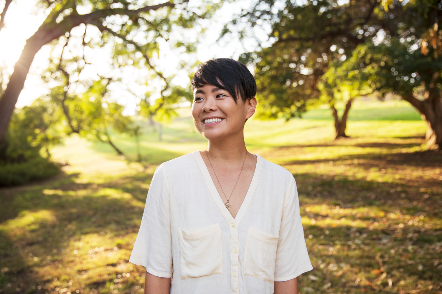 traci ishigo stands in a park surrounded by trees and sunlight, wearing warm smile